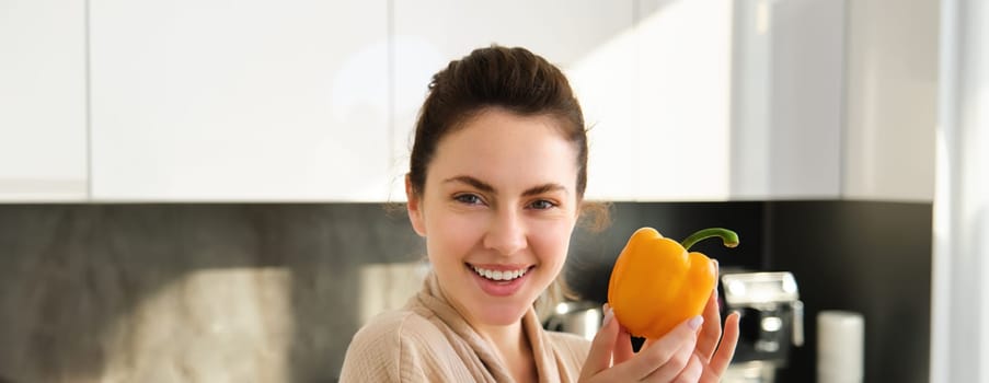 Close up of brunette woman in robe, holding yellow pepper, thinking what to cook, preparing salad or soup, making vegetarian meal with vegetables, standing in kitchen.