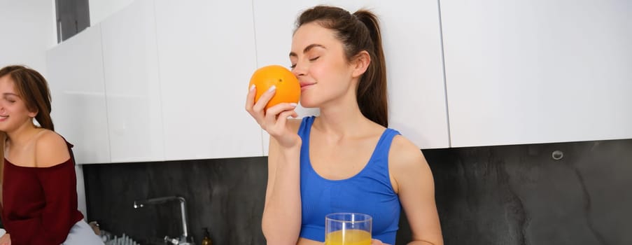 Portrait of fitness instructor, smiling woman, enjoys smell of fresh fruit, drinking orange juice and looking happy, standing in kitchen in activewear.