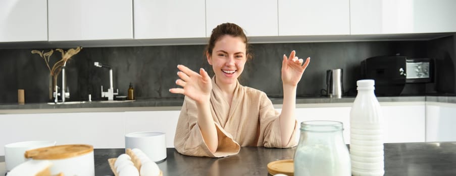 Attractive young cheerful girl baking at the kitchen, making dough, holding recipe book, having ideas.