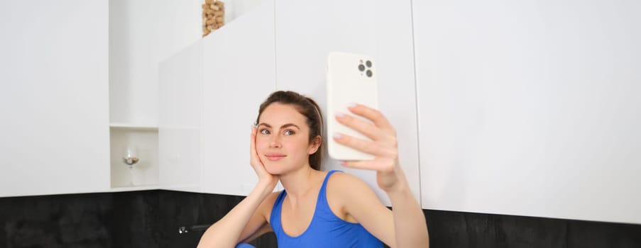 Portrait of fitness girl posing for photo, taking selfie on smartphone app, sitting in kitchen, wearing activewear.