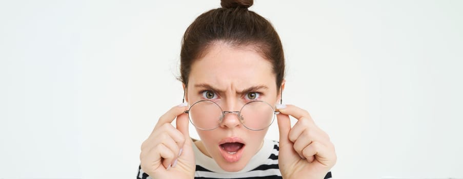 Close up of shocked, insulted young woman, frowning and looking offended, reading smth offensive and upsetting, takes off glasses, stands over white background.