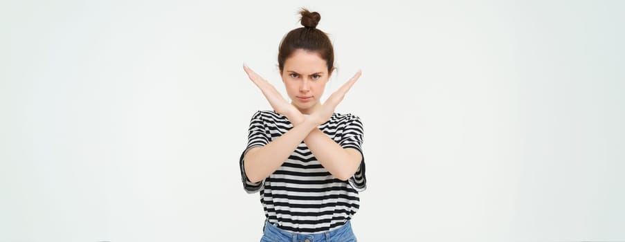Portrait of young serious woman, shows cross, stop gesture, prohibit something, expresses her disapproval, stands over white background.