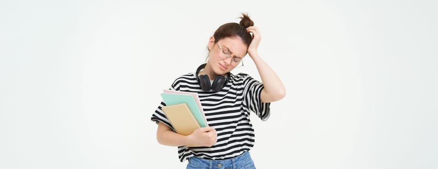 Disappointed young woman, looking tired or upset, wearing glasses, holding study material, documents and notebooks, stands over white background.