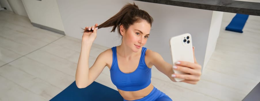 Portrait of beautiful female athlete, woman doing workout at home on rubber yoga mat, takes selfie on smartphone, makes photos for social media of fitness instructor.