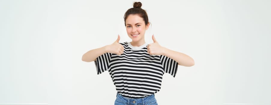 Portrait of cute girl, student shows thumbs up, recommends product, approves choice, likes item, stands over white background.