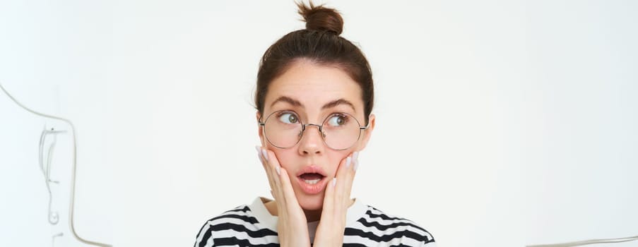 Image of shocked woman in glasses, holds hands on face, gasps and looks speechless, amazed by news, stands over white background.