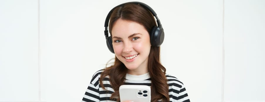 Image of brunette young woman, smiling, listening to music in headphones, watching videos on mobile phone app, holding smartphone, standing over white background.