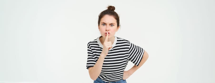 Angry woman shushing, hush at you, press finger to lips, tells to keep quiet, arguing, scolding someone, standing over white background.