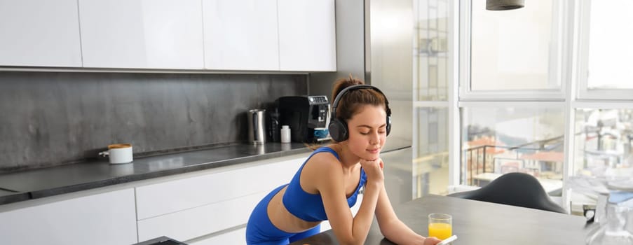 Portrait of young modern woman, beautiful girl in fitness clothes, standing in kitchen, watching yoga video class on smartphone, listening in wireless headphones, drinking juice.