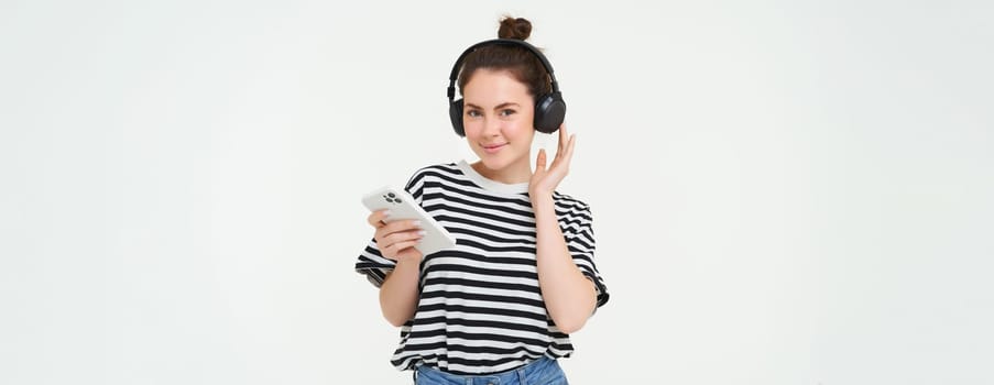 Image of beautiful, modern girl in headphones, listens music, holds mobile phone, changes song, smiling, standing over white background.