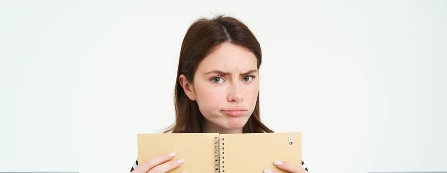 Portrait of girl looking puzzpled, perplxed face of woman, holding notebook, confused by something, standing over white background.