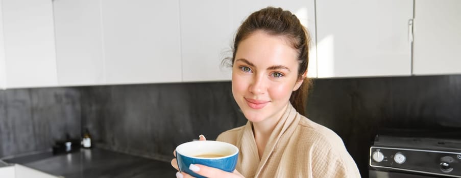 Good-looking young woman with cup of coffee, posing in the kitchen, smiling at camera, enjoys her morning at home, wearing cosy bathrobe.