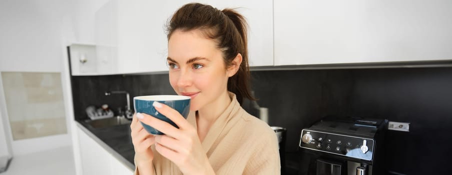 Portrait of beautiful young woman, drinking coffee, freshly made cappuccino, smiling pleased, enjoying her morning, standing in the kitchen.
