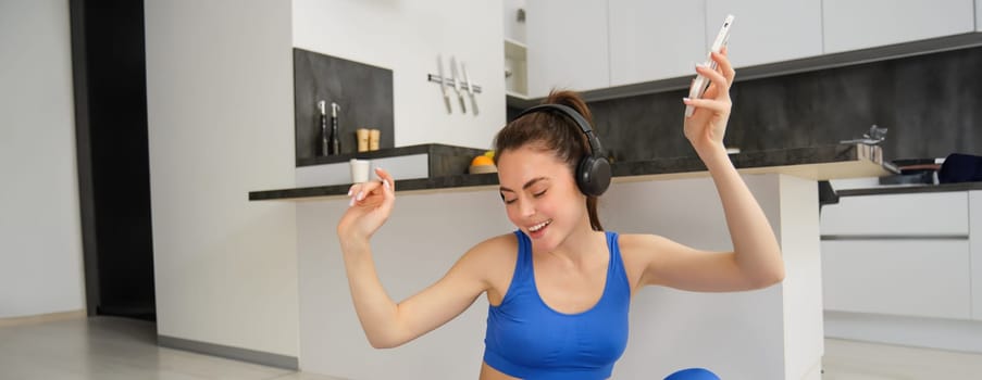 Happy fitness woman dancing, having fun during workout at home, wears headphones, holds smartphone, listens music during exercises.
