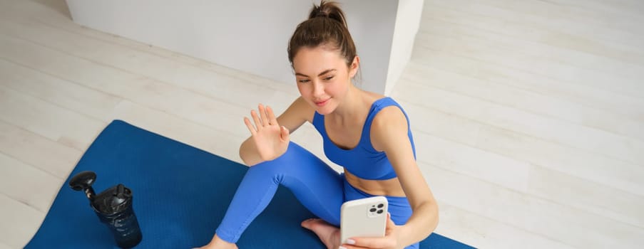 Image of beautiful, atheltic girl sits on yoga mat at home with smartphone, takes selfie and workout, does fitness exercises.