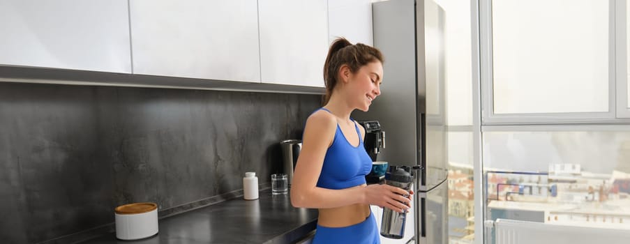 Image of young fitness instructor, woman in sportsbra and leggings, holding water bottle, drinking after workout, standing in kitchen.