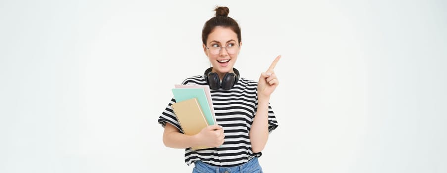 Image of young modern girl, student or teacher in glasses, holding documents and notebooks, pointing at upper left corner with pleased smile, showing advertisement, white background.