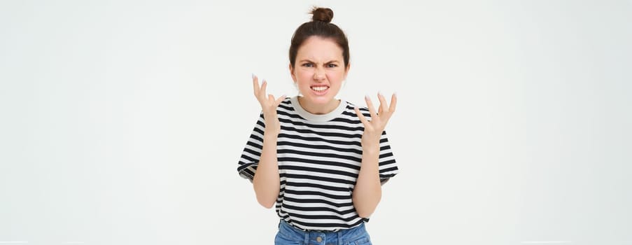 Image of angry woman shouts and shakes hands, stands in casual clothes over white background. Copy space