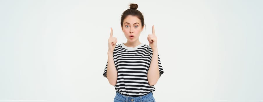 Emotion of amazement. Young woman with impressed face, says wow, points fingers up, shows promo banner on top, recommends product, white background.