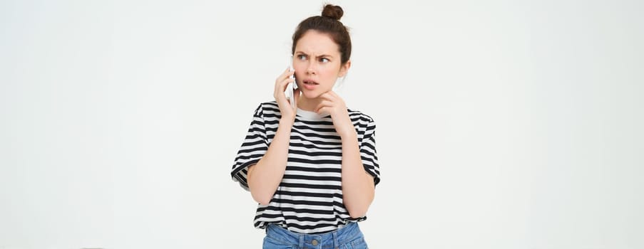 Image of woman with shocked face, looks confused while chats over the phone, listens to person on telephone, white background.