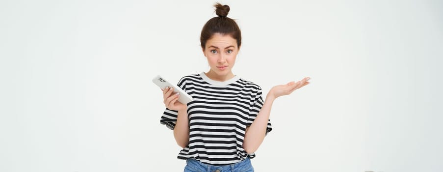 Portrait of shocked girl shrugs shoulders, looks confused reaction to something she saw on mobile phone, stands over white background.