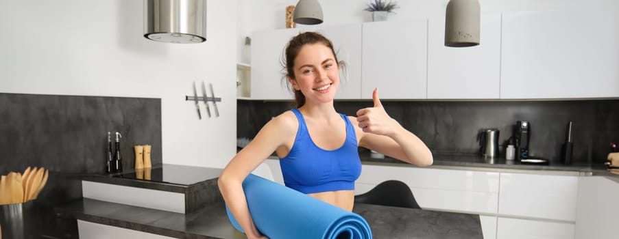 Beautiful young woman, shows thumb up, holds rubber yoga mat, doing fitness exercises from home, gym in living room, recommends fitness exercises.