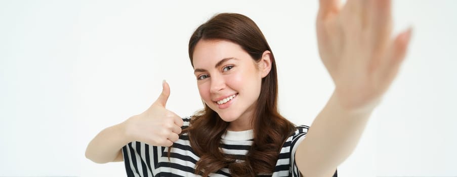 Portrait of girl takes selfie with thumbs up next to something she recommends, likes and approves, stands over white background.