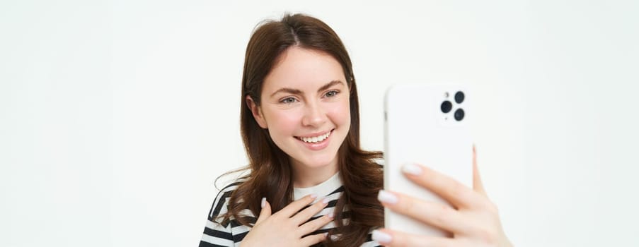 Portrait of happy woman taking photo, laughing while shooting something on smartphone, standing isolated over white background.