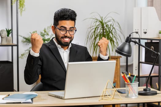 Happy Indian business man working on laptop at home office desk typing browsing celebrating success victory, winning lottery jackpot goal achievement play game good positive news, triumph