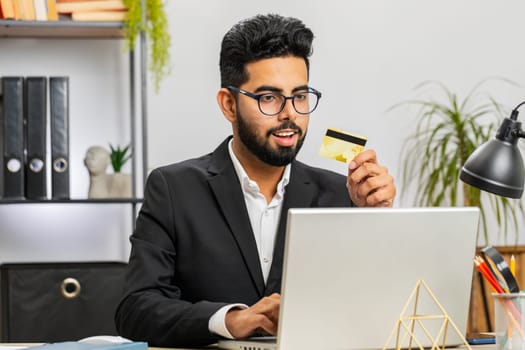 Indian businessman programmer software developer working making online purchase payment shopping with credit bank card and use laptop at home office desk with laptop. Arabian freelancer business man