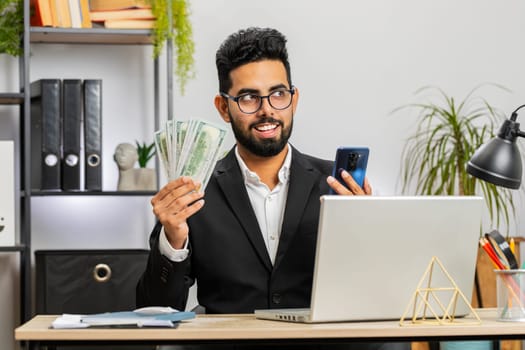 Happy rich Indian bearded businessman counting money dollar cash, use smartphone calculator app at home office workplace. Professional freelancer man rejoicing win, success contract, earning profit