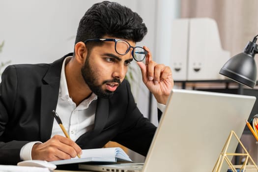 Focused shocked Indian businessman freelancer working at modern home office workplace desk writing notes ideas in notebook. Male Hindu in formal suit preparing planning presentation startup project