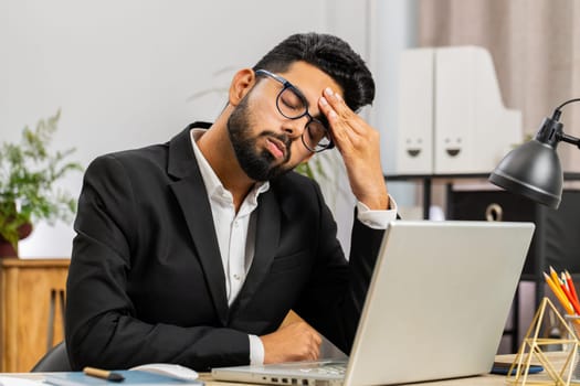 Tired ill Indian businessman suffering from headache problem tension and migraine, stress at home office desk workplace. Bearded freelancer broker worker man works on laptop notebook. Overworking