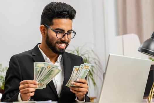 Rich Indian businessman working on laptop pc counting money cash, calculate earnings income profit at home office workplace. Happy professional freelancer man. Business people. Employment occupation