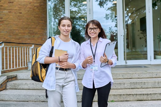 Female teacher, mentor, coach and teenage girl student with backpack together, looking at camera outdoor, school building background. Adolescence, education, knowledge, communication