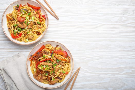 Two bowls with Chow Mein or Lo Mein, traditional Chinese stir fry noodles with meat and vegetables, served with chopsticks top view on rustic white wooden background table, space for text.