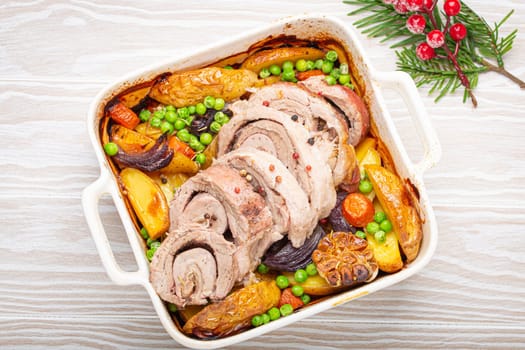 Festive Christmas rolled sliced pork roasted in white casserole dish with potatoes, vegetables and herbs on rustic white wooden background top view. Baked pork roll with vegetables for Xmas dinner.