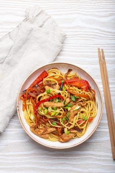 White bowl with Chow Mein or Lo Mein, traditional Chinese stir fry noodles with meat and vegetables, served with chopsticks top view on rustic white wooden background table.