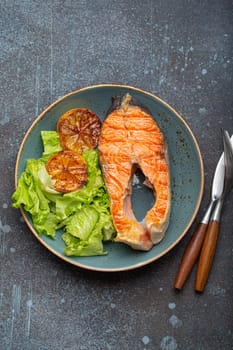 Grilled fish salmon steak and green salad with lemon on ceramic plate on rustic blue stone background top view, balanced diet or healthy nutrition meal with salmon and veggies.