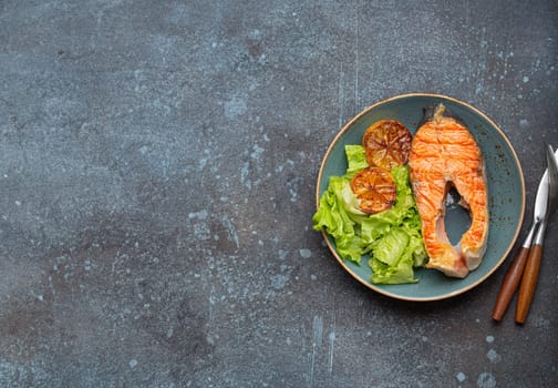 Grilled fish salmon steak and green salad with lemon on ceramic plate on rustic blue stone background top view, balanced diet or healthy nutrition meal with salmon and veggies, space for text.