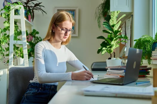 Teenage female student sitting at home at desk, writing in notebook using laptop computer. Girl studies online, remote lessons, prepares for tests, watches educational webinars. Technology adolescence