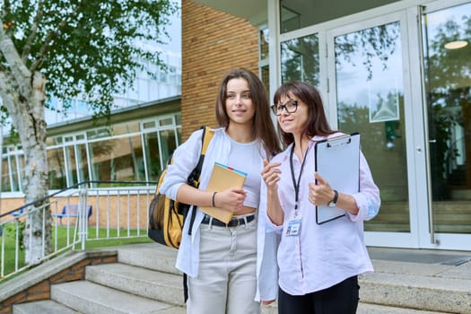 Teenage girl high school student with backpack talking to female teacher, mentor, coach, standing outdoors on steps of educational building. Adolescence, education, knowledge, communication