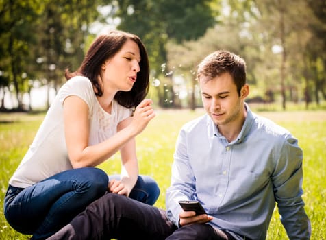 Young woman blowing dandelion to face of her boyfriend holding mobile phone