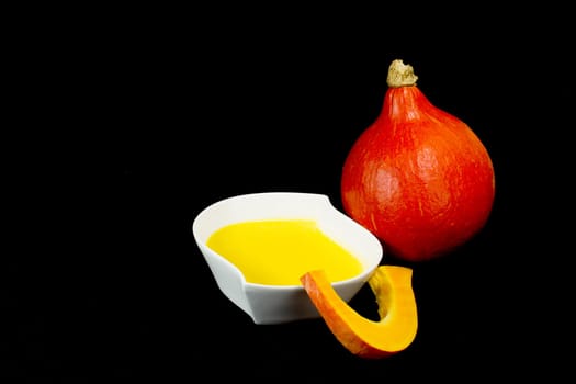 White bowl of pumpkin soup with cream. One piece of pumpkin is leaning on a bowl. Red pumpkin is in the background. All isolated on black background.