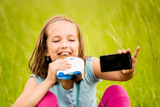 Child is taking photo with mobile phone camera while eating chocolate outdoor in nature