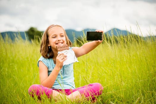 Child is taking photo with mobile phone camera while eating chocolate outdoor in nature