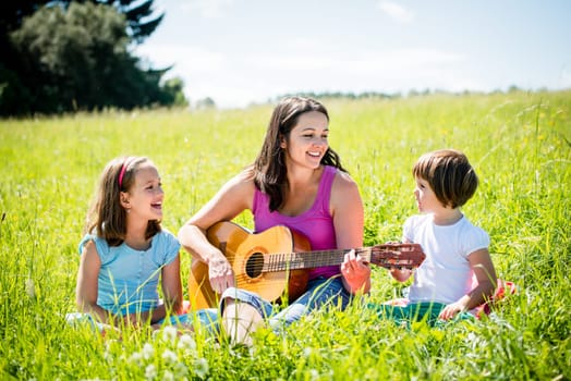 Woman playing guitar to her children in nature on sunny summer day