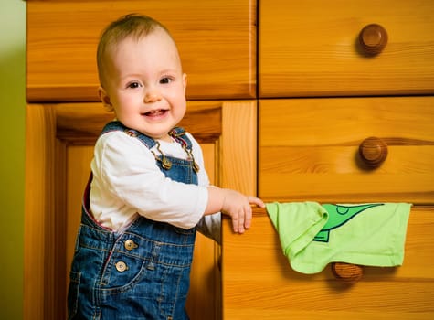 Baby opening drawer with clothes on wooden furniture at home