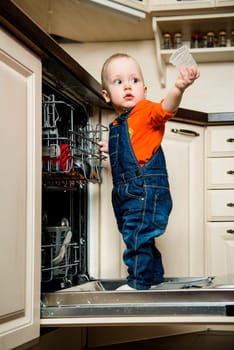 Cute baby standing on dishwasher in kitchen and helps it unload