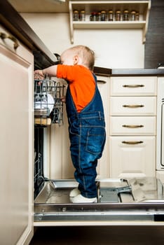 Cute baby standing on dishwasher in kitchen and helps it unload